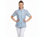 Modelo de medio cuerpo luciendo una blusa médica de color azul claro con estampado de personajes animados (Snoopy/Disney). La blusa tiene cuello Mao, manga corta y cierre de cremallera central. Se muestra combinada con un pantalón blanco sobre un fondo blanco limpio.