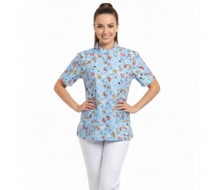 Modelo de medio cuerpo luciendo una blusa médica de color azul claro con estampado de personajes animados (Snoopy/Disney). La blusa tiene cuello Mao, manga corta y cierre de cremallera central. Se muestra combinada con un pantalón blanco sobre un fondo blanco limpio.