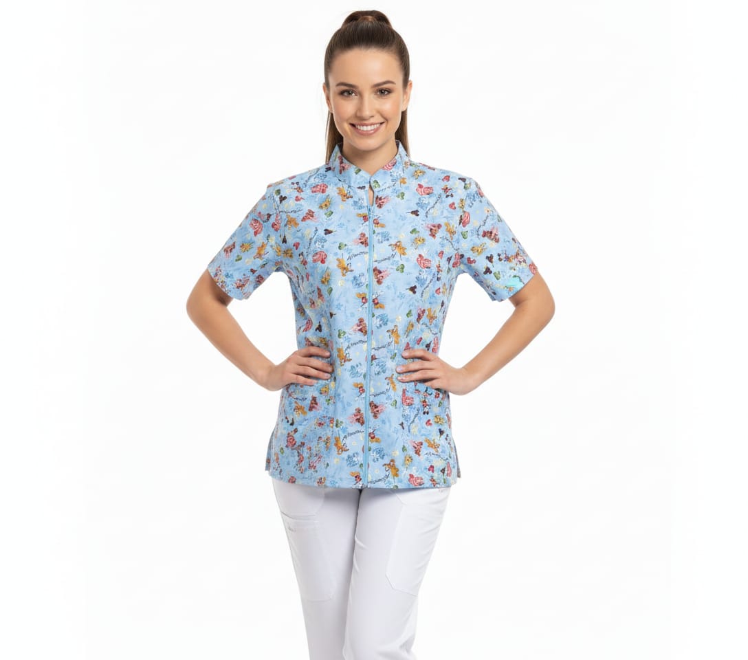 Modelo de medio cuerpo luciendo una blusa médica de color azul claro con estampado de personajes animados (Snoopy/Disney). La blusa tiene cuello Mao, manga corta y cierre de cremallera central. Se muestra combinada con un pantalón blanco sobre un fondo blanco limpio.